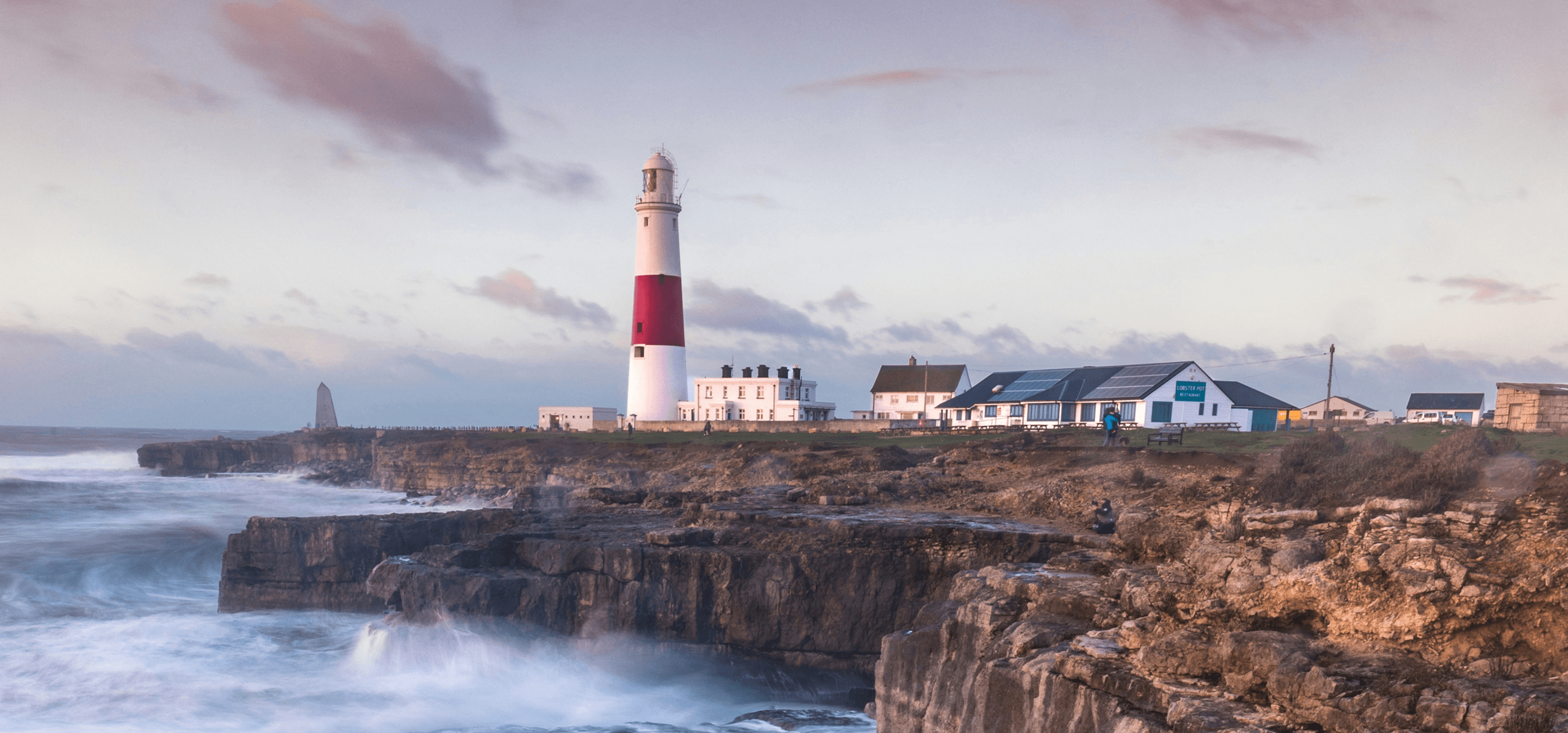 Wellen brechen auf einer britischen Klippe mit einem Leuchtturm in der Ferne