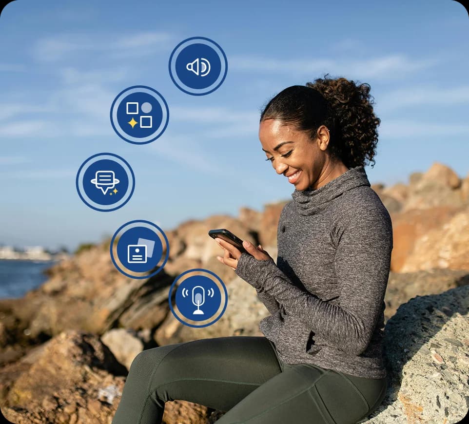 Eine Frau sitzt draußen auf Felsen am Wasser, lächelt und schaut auf ihr Handy, umgeben von Icons der Rosetta Stone Sapphire-Funktionen.